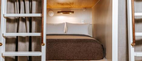 Un espacio para dormir compacto y bien iluminado con cama, cortinas y una pequeña estantería.