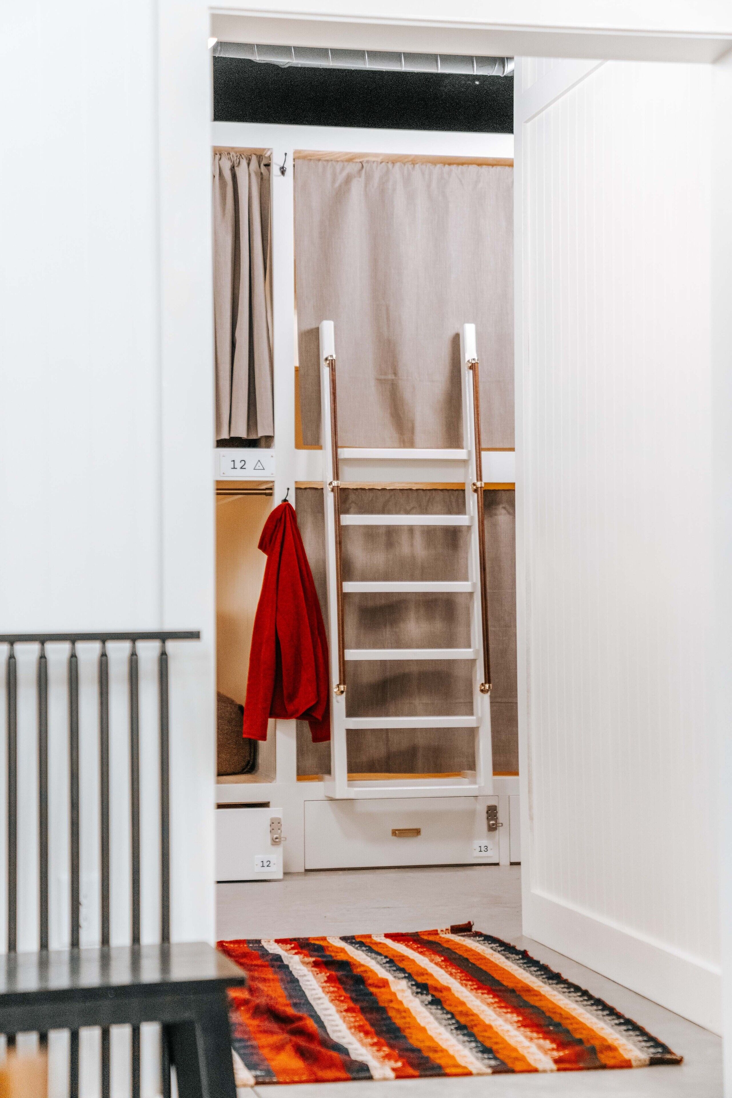 Une pièce avec un lit superposé, une échelle, une veste rouge accrochée au mur et un tapis à rayures sur le sol.