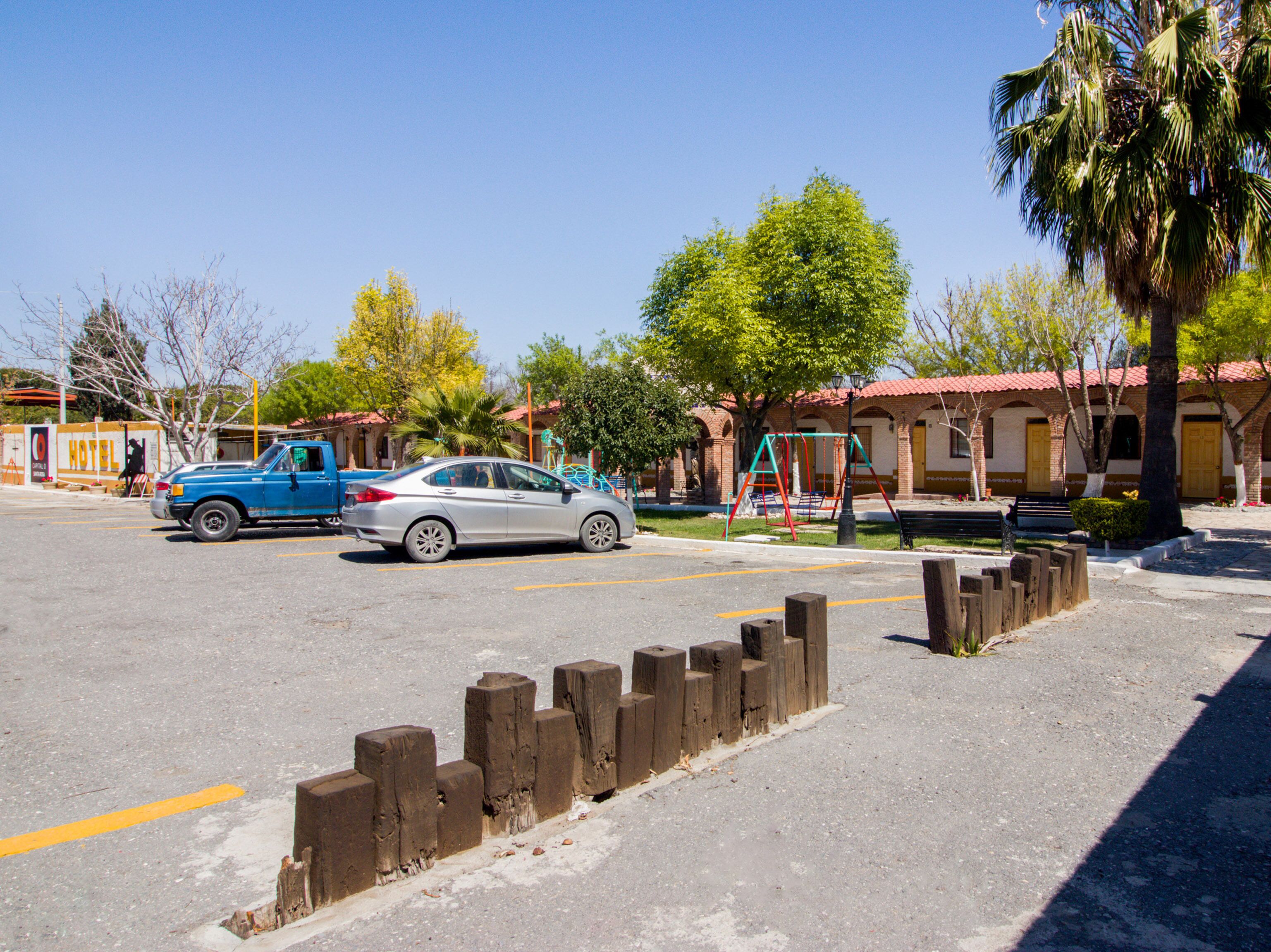 A hotel with a red-tiled roof, a playground, and a parking lot with a blue truck and a silver car.