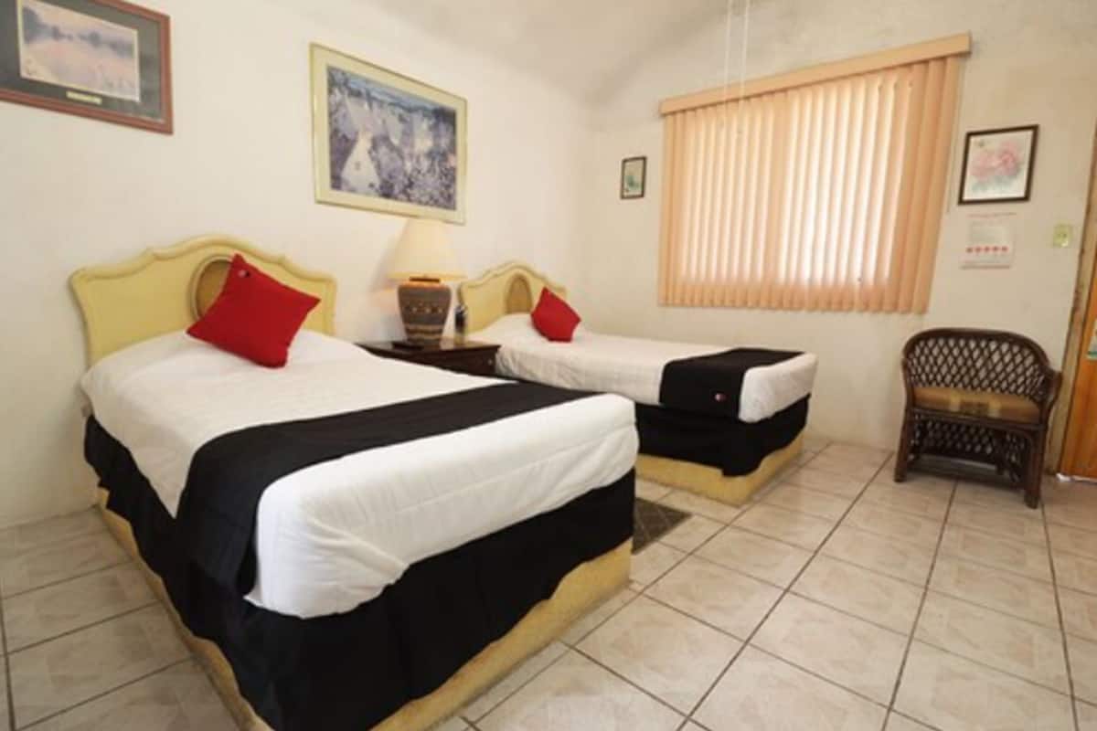 A room with two beds, each with a red pillow, a nightstand with a lamp, a wicker chair, and a window with blinds.