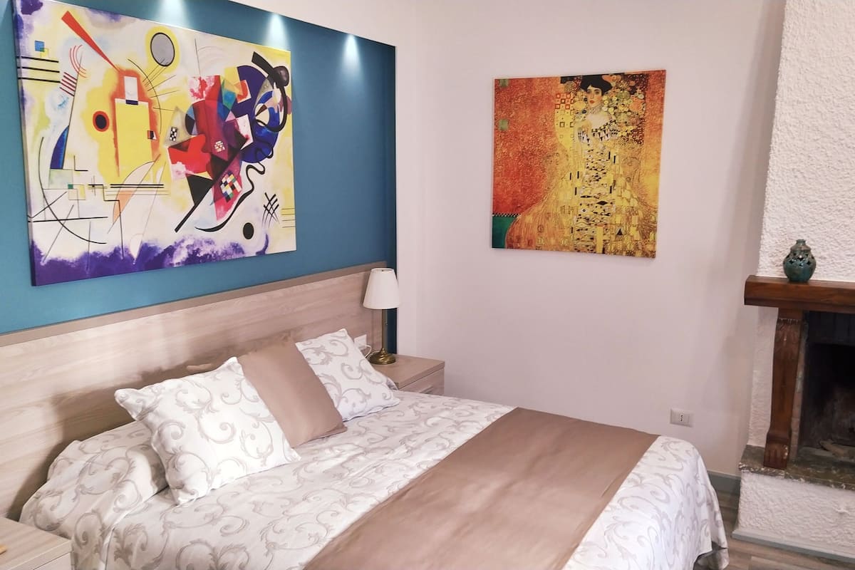 A bedroom with a bed, a nightstand, a lamp, a fireplace, and two paintings on the wall.