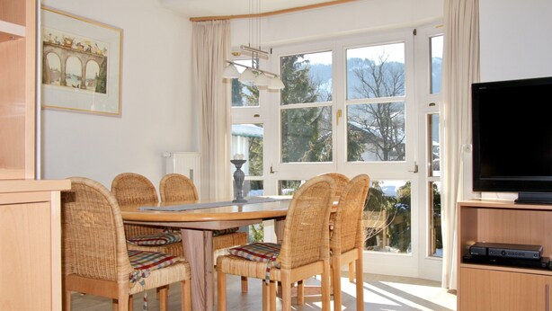 Uma sala de jantar com mesa de madeira e cadeiras de vime, uma televisão sobre suporte de madeira, e uma janela com vista para árvores e montanhas.
