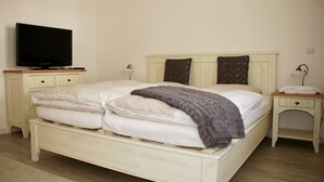 Uma cama bem arrumada com travesseiros e cobertor, uma televisão em um suporte, uma mesa de cabeceira com abajur e um criado-mudo com luminária.