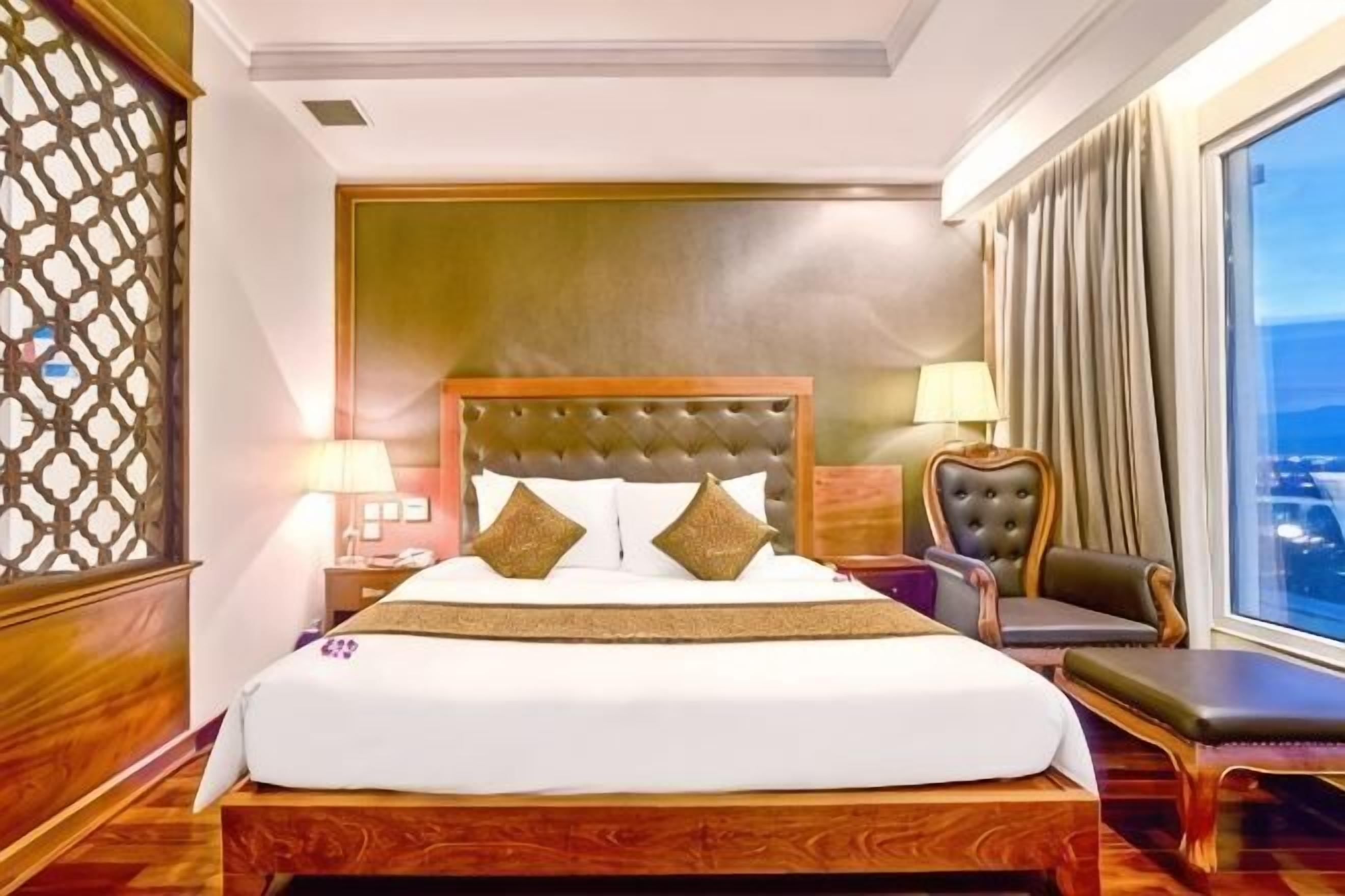 Une chambre d’hôtel avec un grand lit, une tête de lit en bois, une table de chevet avec une lampe, une chaise et une vue sur la ville.