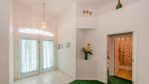 Una habitación con alfombra verde, paredes blancas, una araña y una puerta con cortina, además de un jarrón pequeño con flores.