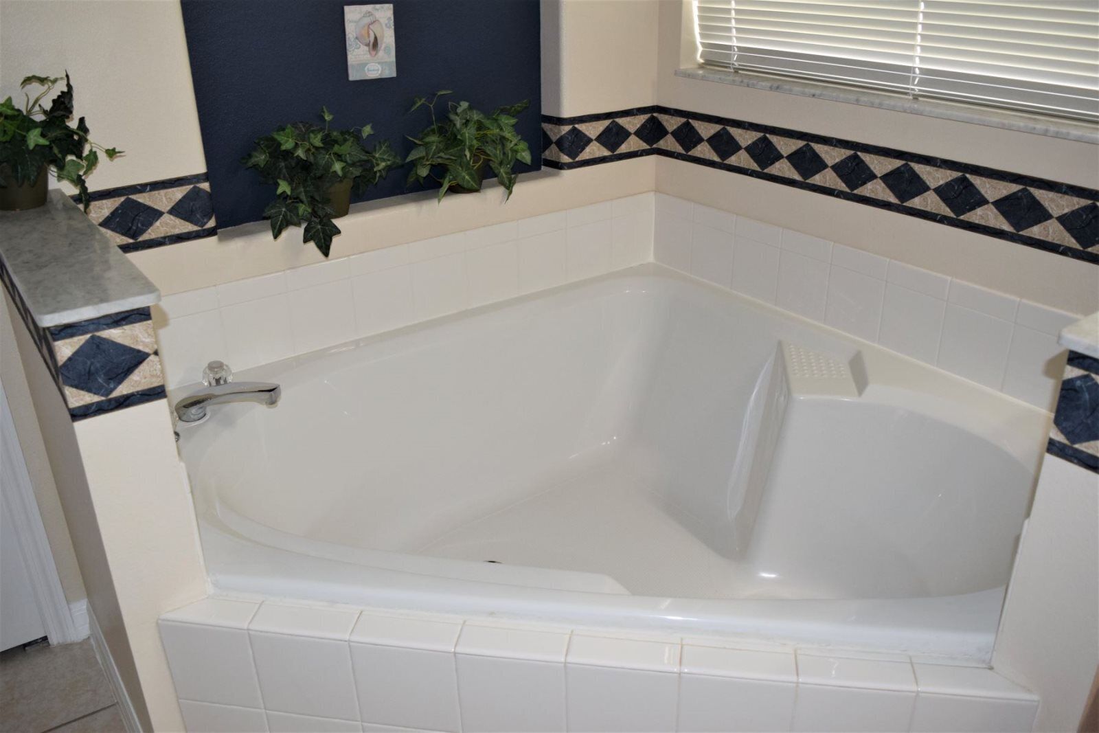 Une salle de bain avec une baignoire blanche, une douche et un sol carrelé. Des plantes en pot sont disposées sur le mur, et il y a une fenêtre avec des stores.
