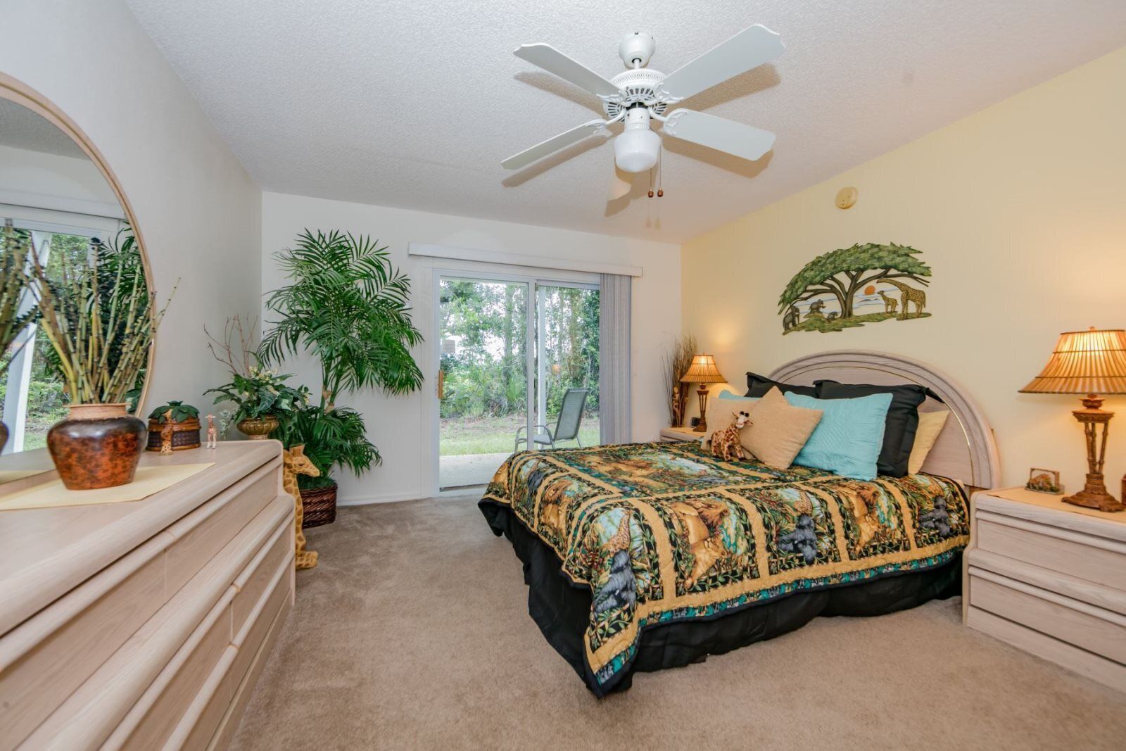 A bedroom with a bed, a ceiling fan, a large window, and several potted plants.