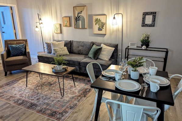 A living room with a sofa, armchair, coffee table, and dining table set for four. There are framed pictures on the wall and a potted plant on the table.