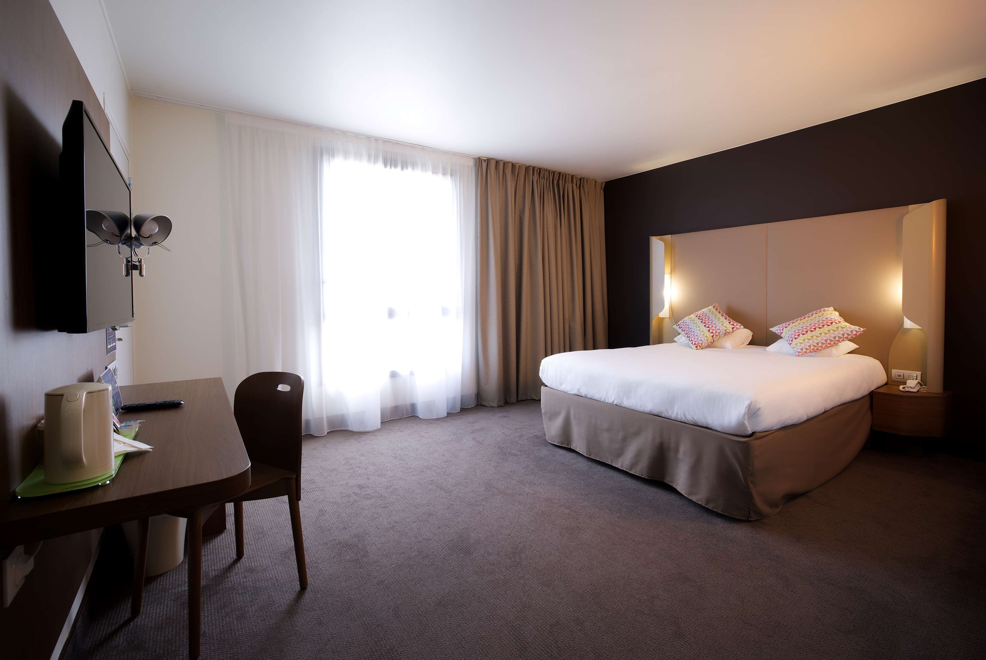 Hotellihuone, jossa on sänky, työpöytä tuolilla, televisio ja ikkuna, jossa on verhot.