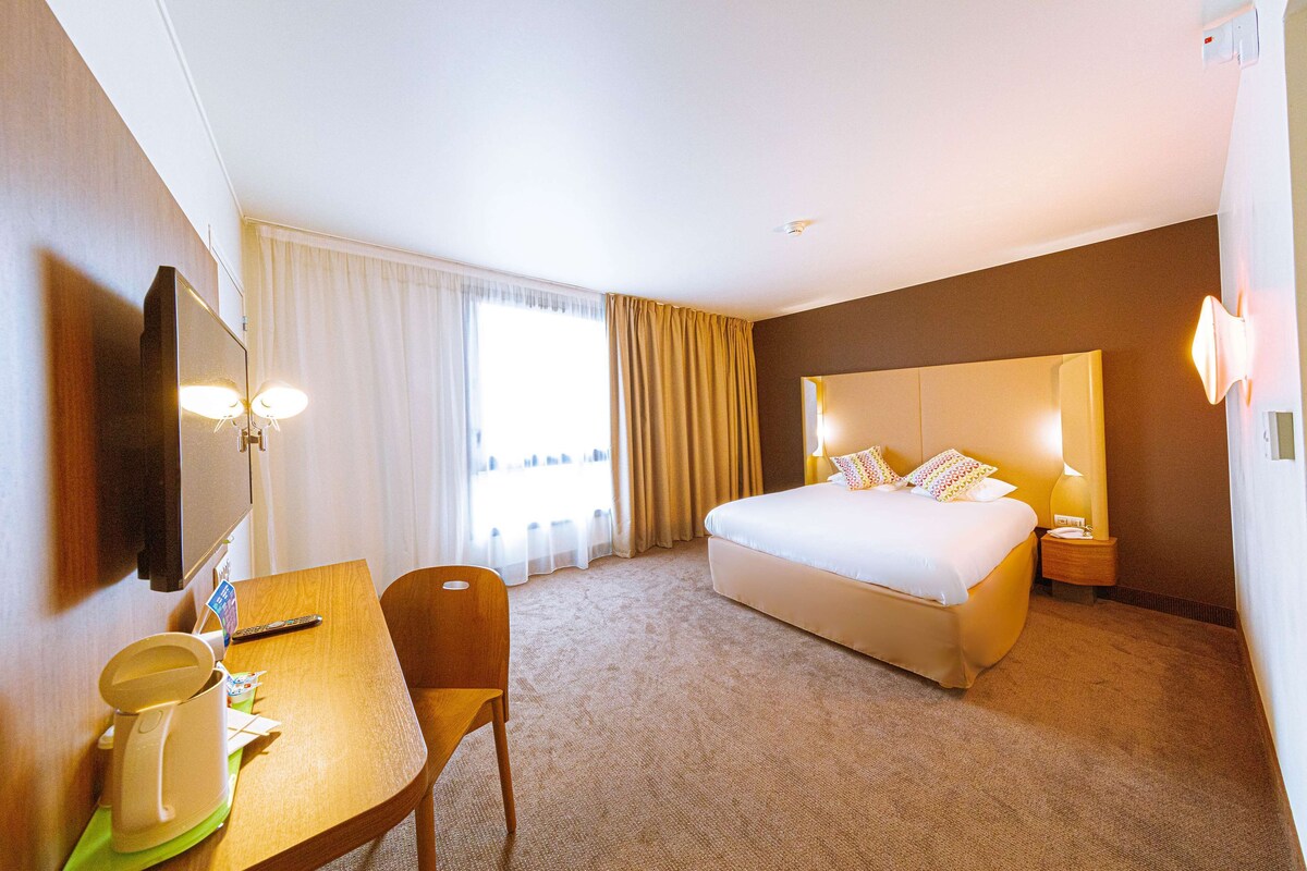 A hotel room with a bed, a desk with a chair, a television, and a window with curtains.