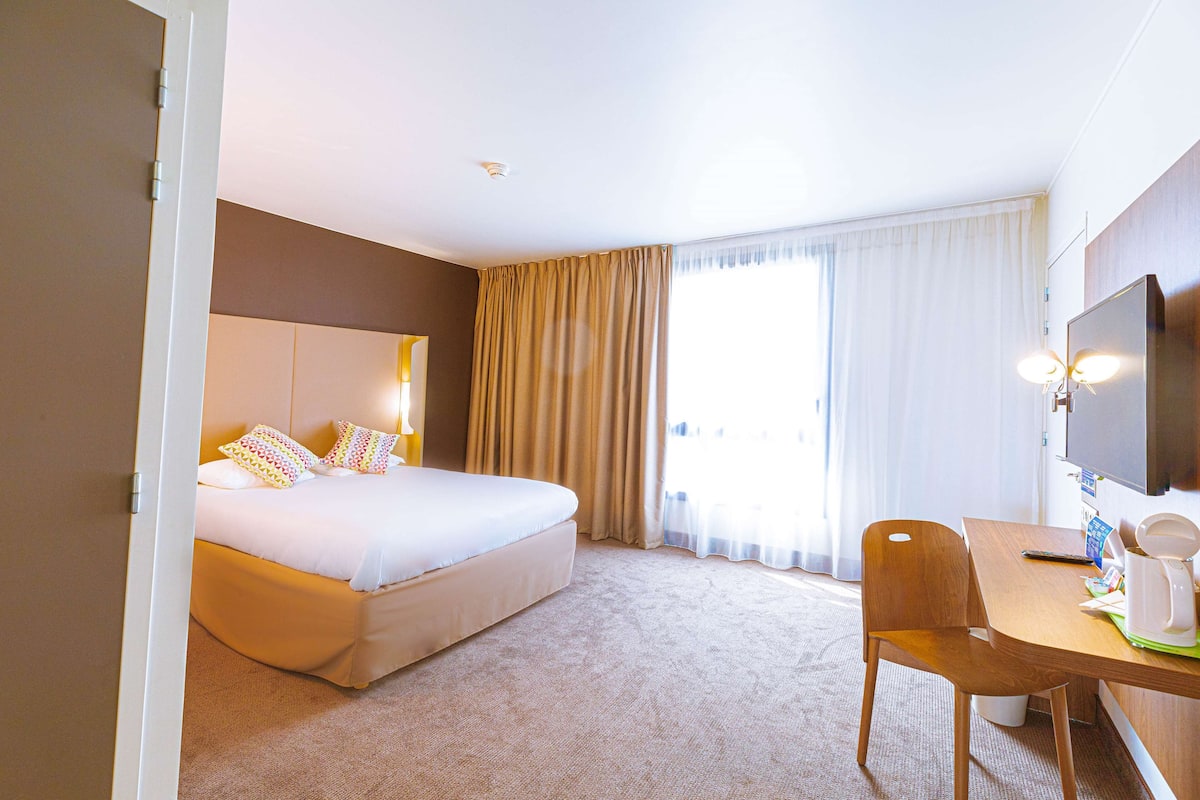 Habitación de hotel con cama, escritorio, silla, televisor y ventana con cortinas.