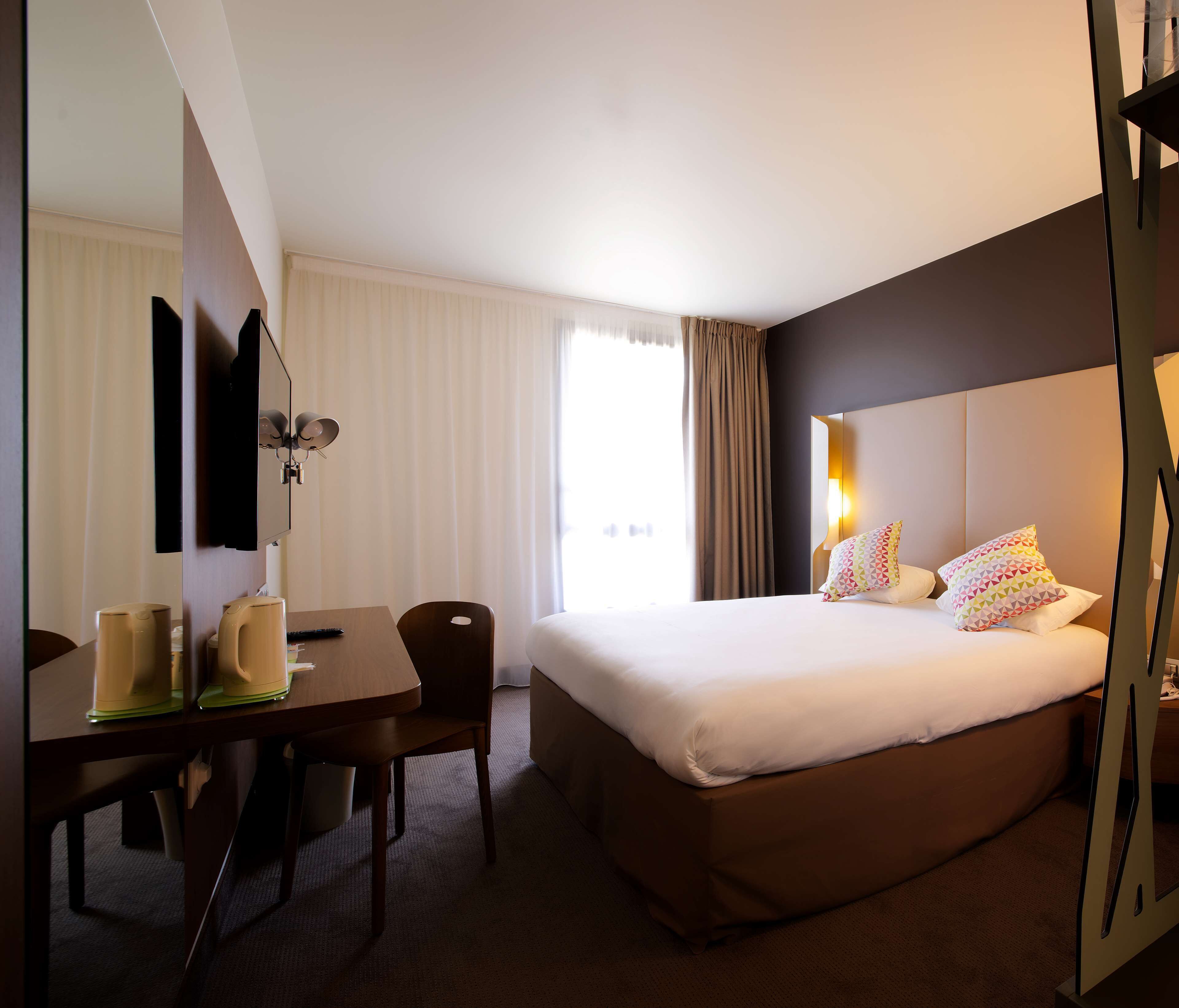 Ein Hotelzimmer mit einem großen Bett, einem Schreibtisch mit zwei Tassen, einem Sessel, einem Flachbildfernseher und einem Fenster mit Vorhängen.