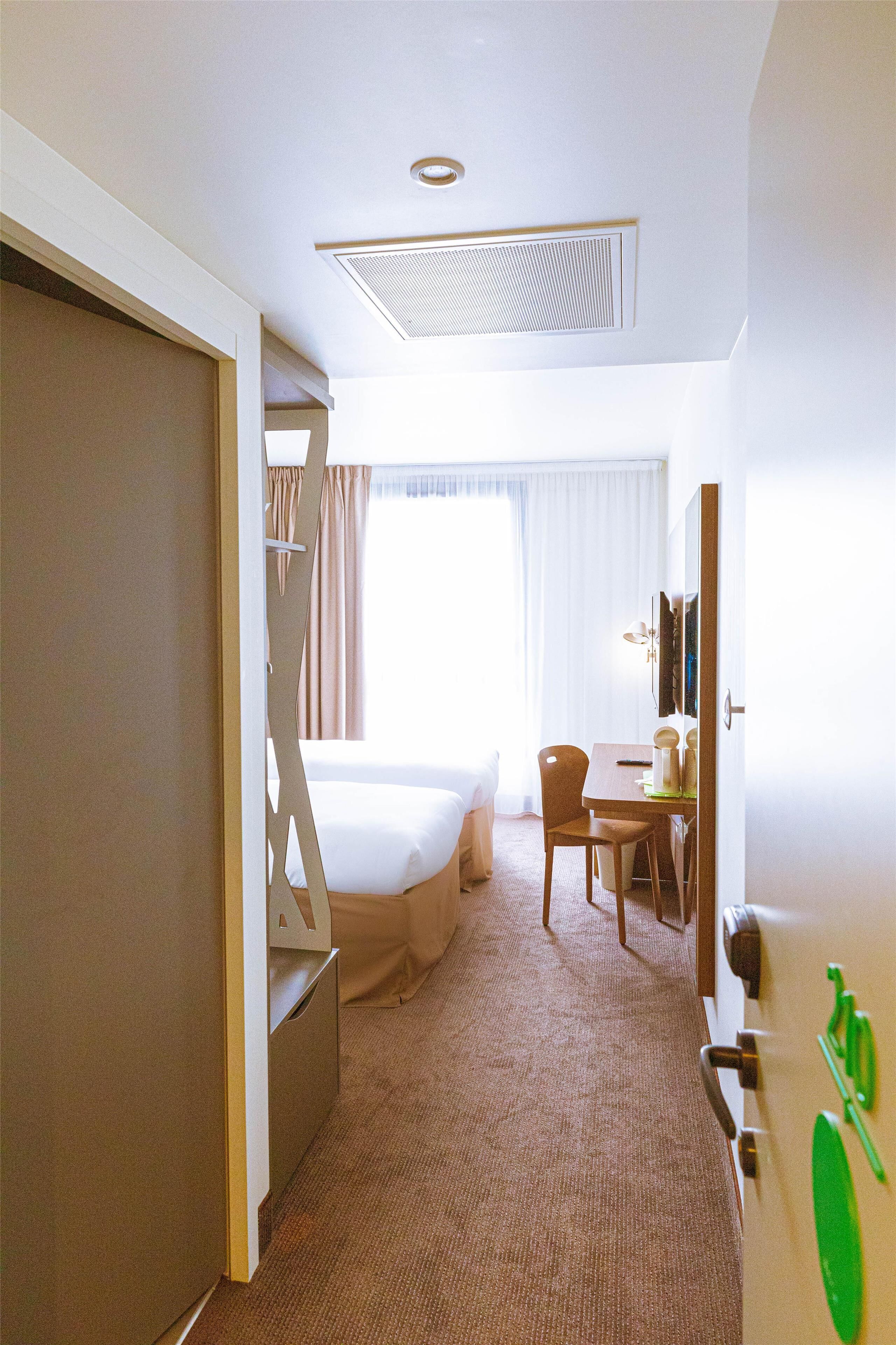 A hotel room with a bed, a desk, and a chair. There is a window with curtains, a door, and a coat rack with a green item on it.