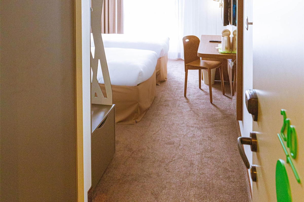 A hotel room with a bed, a desk, and a chair. There is a window with curtains, a door, and a coat rack with a green item on it.
