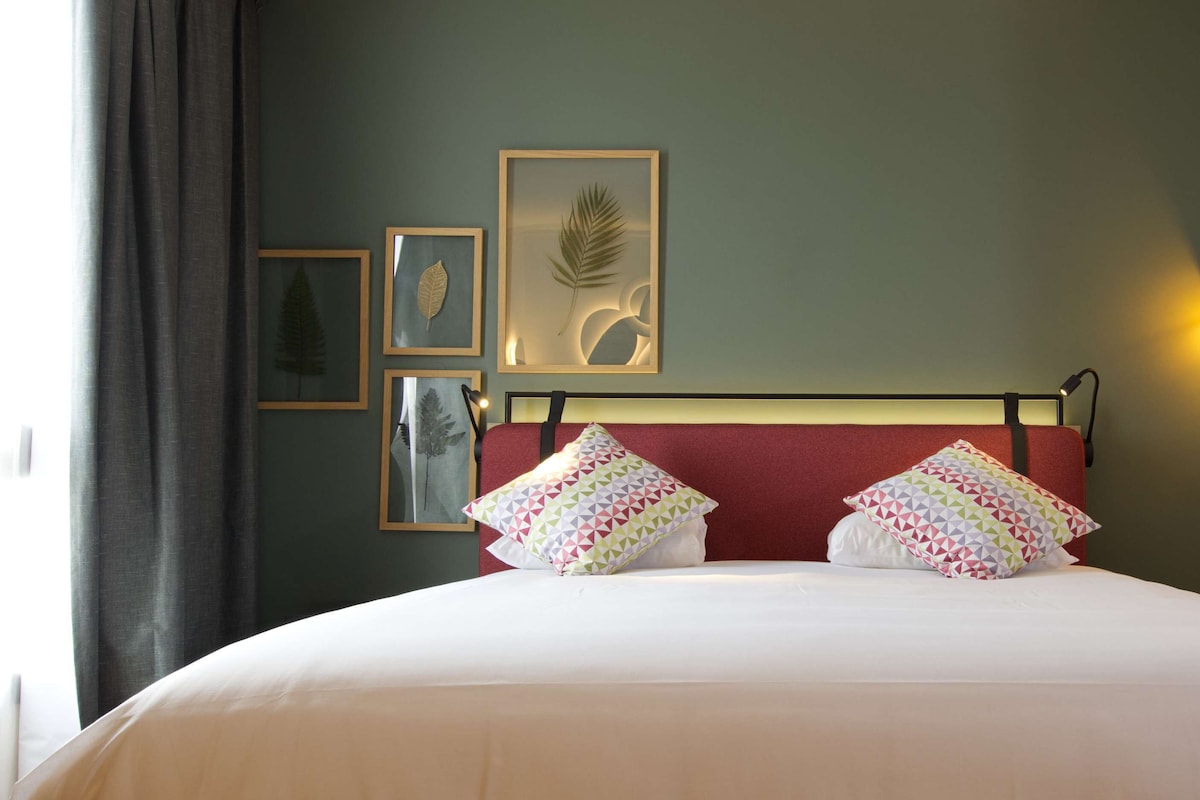 A neatly made bed with white bedding and two patterned pillows, a red headboard, and wall-mounted artwork including leaf designs.