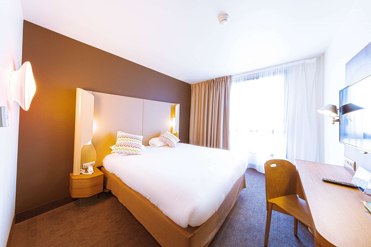 A hotel room with a large bed, a desk, and a chair. There is a window with curtains, a lamp on the nightstand, and a television on the wall.