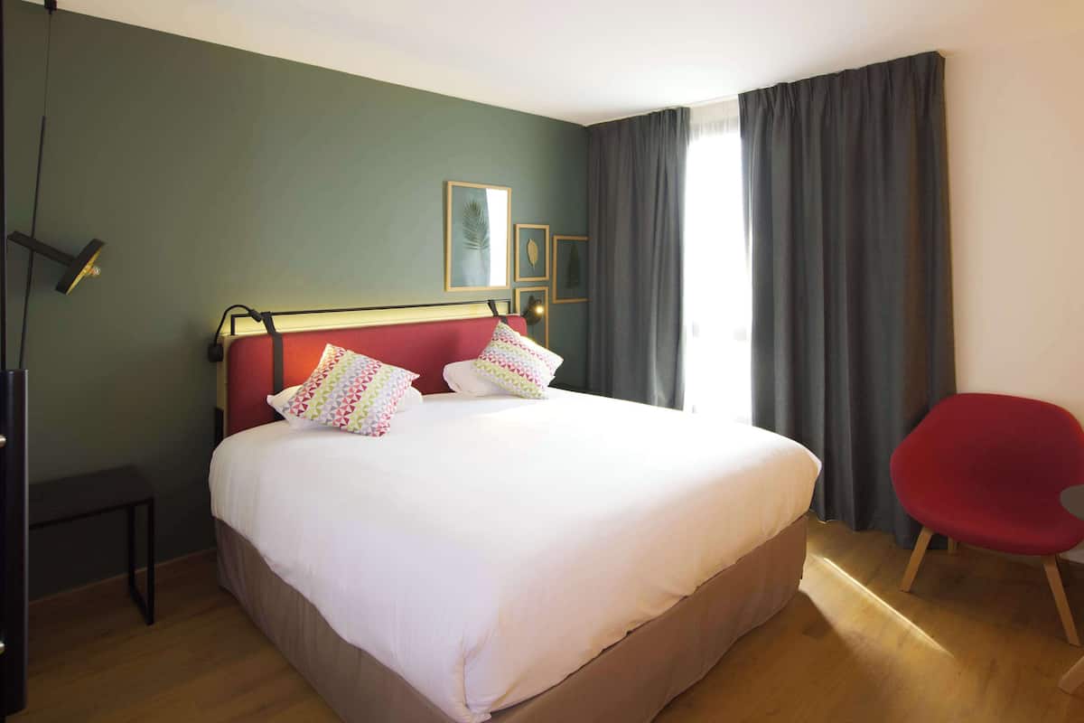 Una cama bien tendida con sábanas blancas y almohadas coloridas, una silla roja y un escritorio con una lámpara.