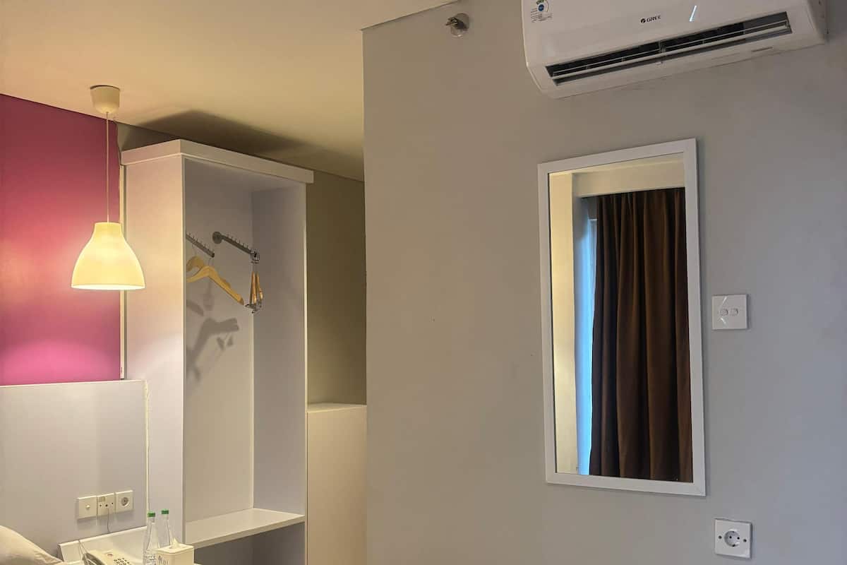 A hotel room with a bed, a bedside table with a lamp, a wardrobe, an air conditioning unit, and a window with curtains.