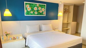 A hotel room with a large bed, white bedding, a blue accent wall, a painting of flowers, a bedside table with a vase of flowers, a phone, and a wardrobe. - Kyriad Haka Makassar (Makassar)