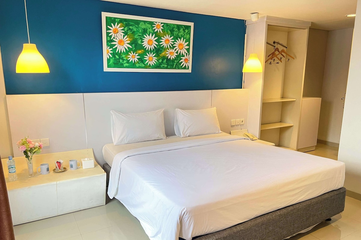 A hotel room with a large bed, white bedding, a blue accent wall, a painting of flowers, a bedside table with a vase of flowers, a phone, and a wardrobe.