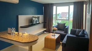 A modern hotel room with a blue accent wall, a flat-screen TV on a black shelf, a wooden coffee table, a dark sofa, and a large window with curtains. - Kyriad Haka Makassar (Makassar)