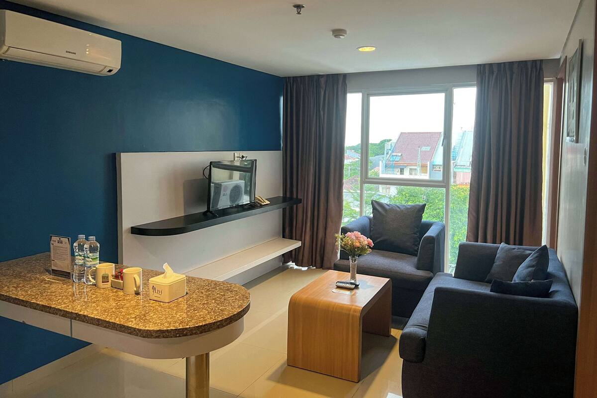 A modern hotel room with a blue accent wall, a flat-screen TV on a black shelf, a wooden coffee table, a dark sofa, and a large window with curtains.