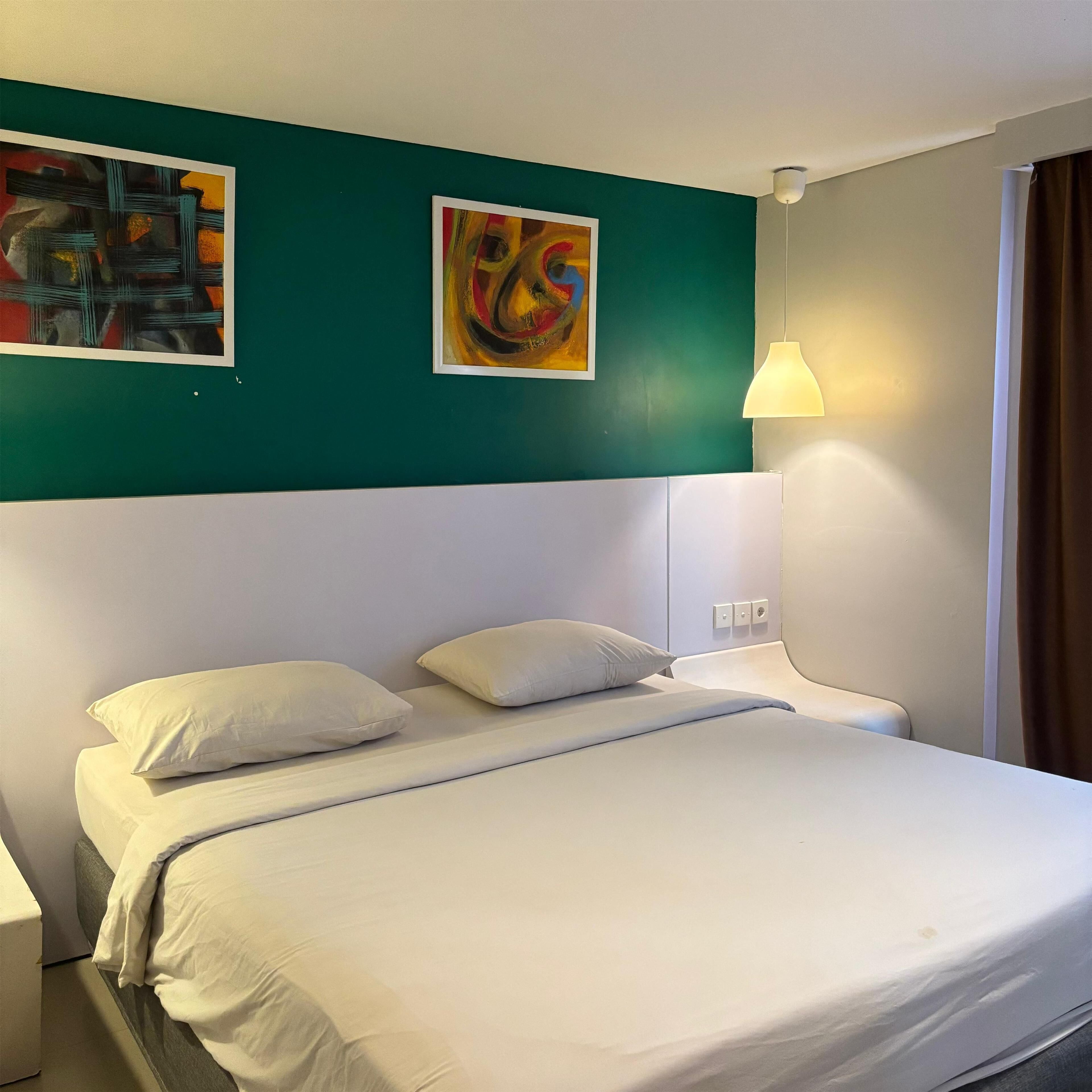 A hotel room with a bed, white bedding, a teal accent wall, and two paintings.