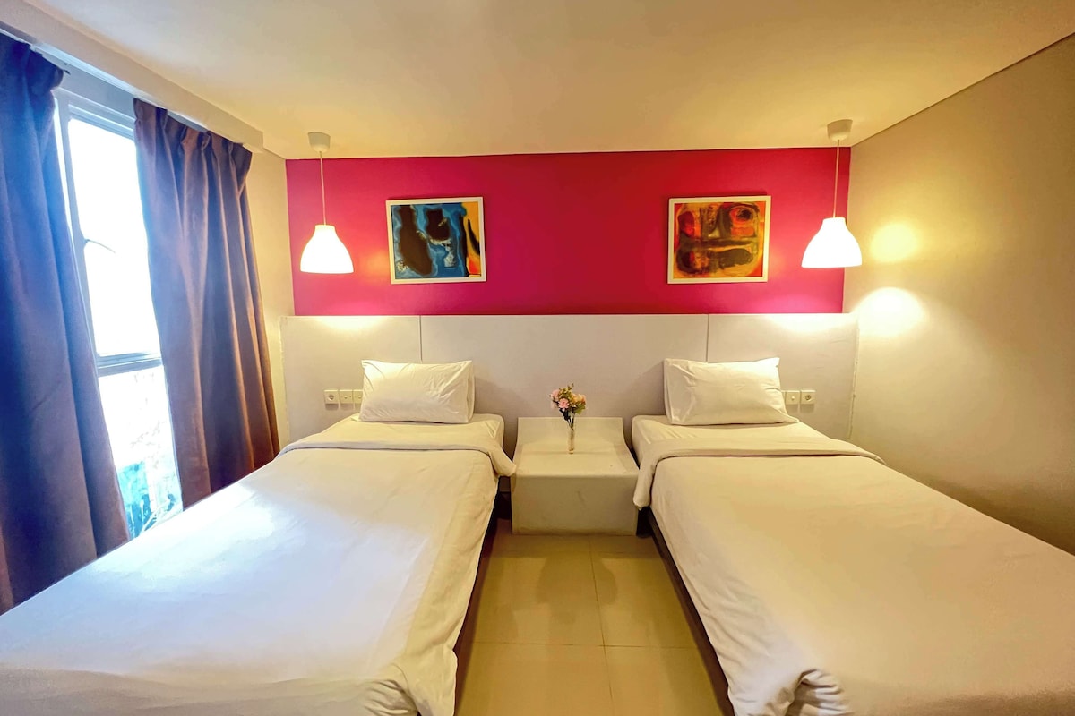 A hotel room with two single beds, a small bedside table with a vase, and a window with curtains.