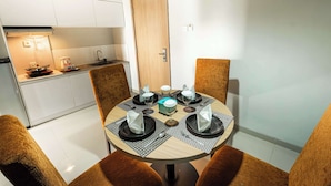 A dining area with a round table set for four, surrounded by brown upholstered chairs. A kitchen with white cabinets and a wooden door is visible in the background. - Kyriad Haka Makassar (Makassar)