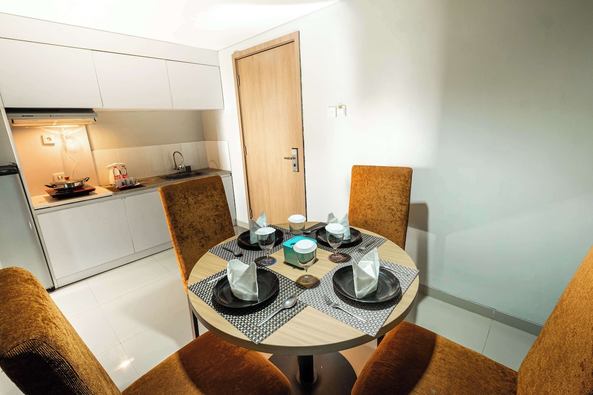 A dining area with a round table set for four, surrounded by brown upholstered chairs. A kitchen with white cabinets and a wooden door is visible in the background.