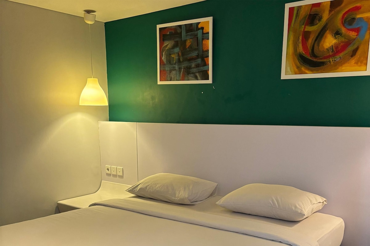 A neatly made bed with white linens in a room with teal walls, a white headboard, and a single bedside lamp.