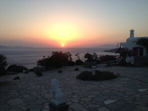 A sunset over the sea with a stone pathway leading to a white building with a statue in the foreground.