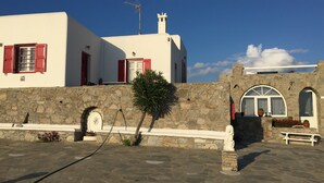 A building with white walls, red shutters, and a stone wall. There is a small tree in front of the stone wall. - Cozy apartment for 2 near Ornos with sea and sunset view  (Ornos Mykonos)