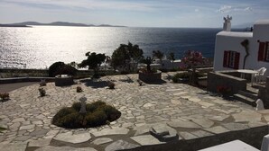 Une cour dallée de pierres, offrant une vue sur la mer, et entourée de bâtiments blancs aux volets rouges.