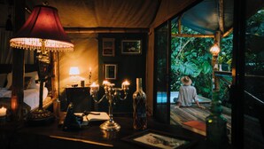 A cozy room with a wooden ceiling, a large window overlooking a lush garden, a table with candles, and a person in a hat outside. - Colina Secreta - Glamping and Villas (Cahuita)