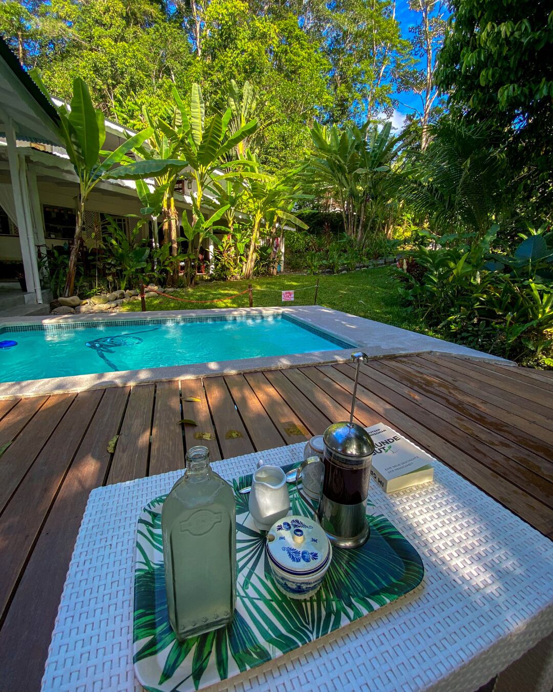 A poolside setting with a wooden deck, a table with a tray holding a bottle, a cup, and a thermos, surrounded by lush greenery and a clear blue pool.