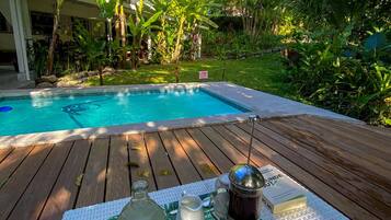 A poolside setting with a wooden deck, a table with a tray holding a bottle, a cup, and a thermos, surrounded by lush greenery and a clear blue pool.