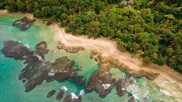 An aerial view of a tropical beach with clear turquoise waters, rocky formations, and dense green foliage. - Colina Secreta - Glamping and Villas (Cahuita)