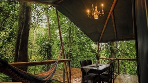 An outdoor wooden deck with a table and chairs, surrounded by lush greenery and trees. - Colina Secreta - Glamping and Villas (Cahuita)
