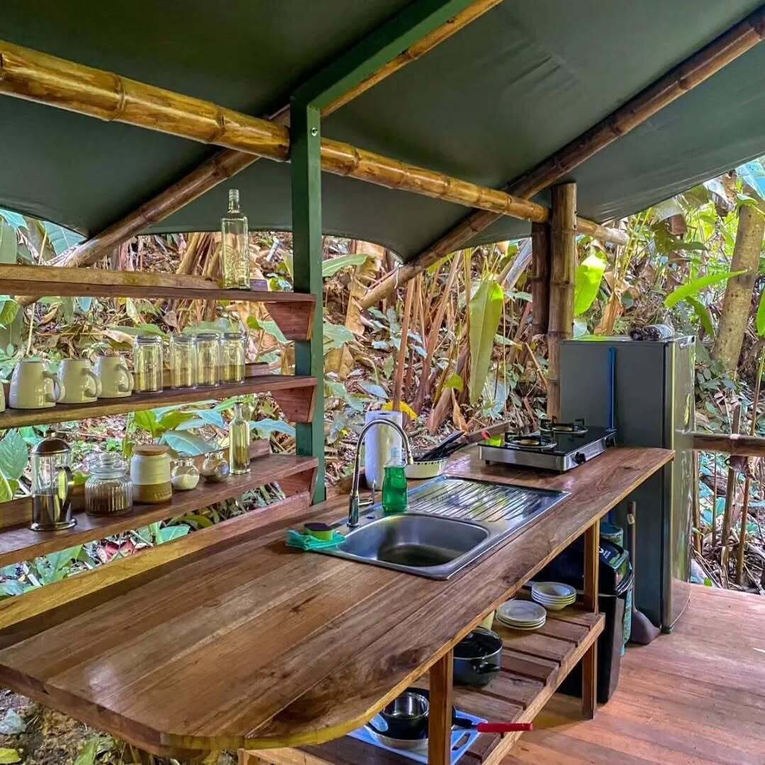 A rustic kitchen with a wooden countertop, shelves with jars, a sink, and a refrigerator.
