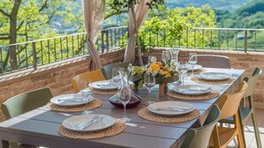 An outdoor dining table set with plates, cutlery, and wine glasses, surrounded by chairs, with a scenic view of trees and mountains in the background.