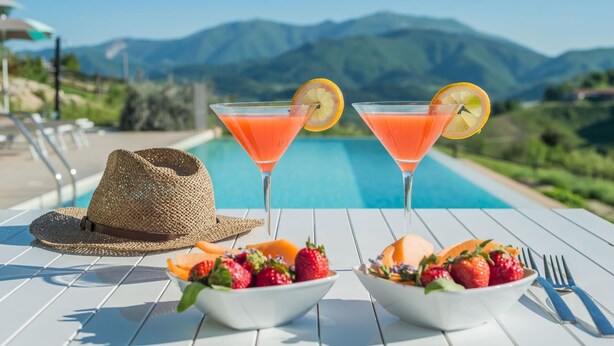 A poolside table with two martini glasses, a straw hat, and bowls of fruit.