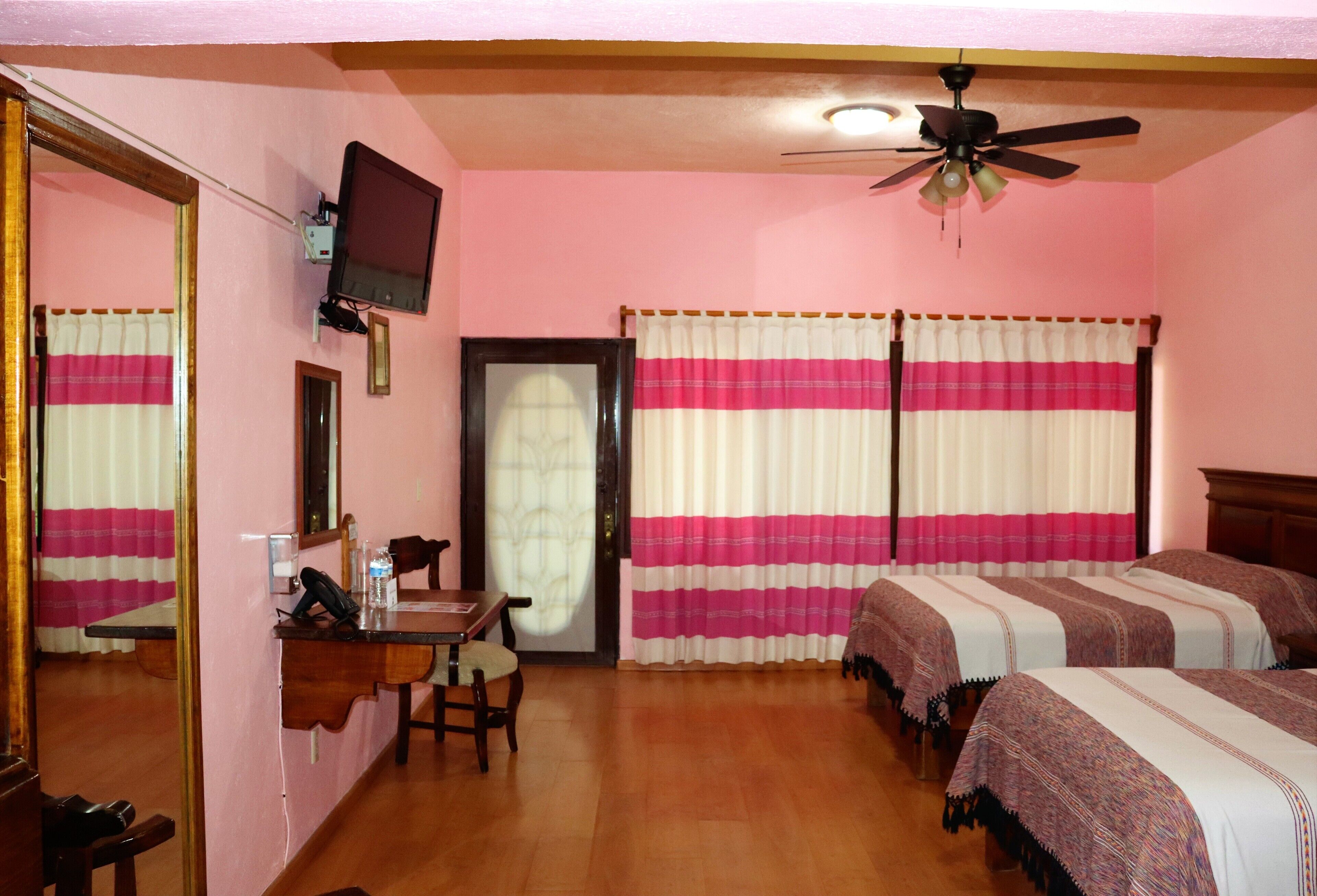 Quarto de hotel com duas camas, uma mesa de trabalho com telefone, uma televisão fixada na parede e um ventilador de teto.