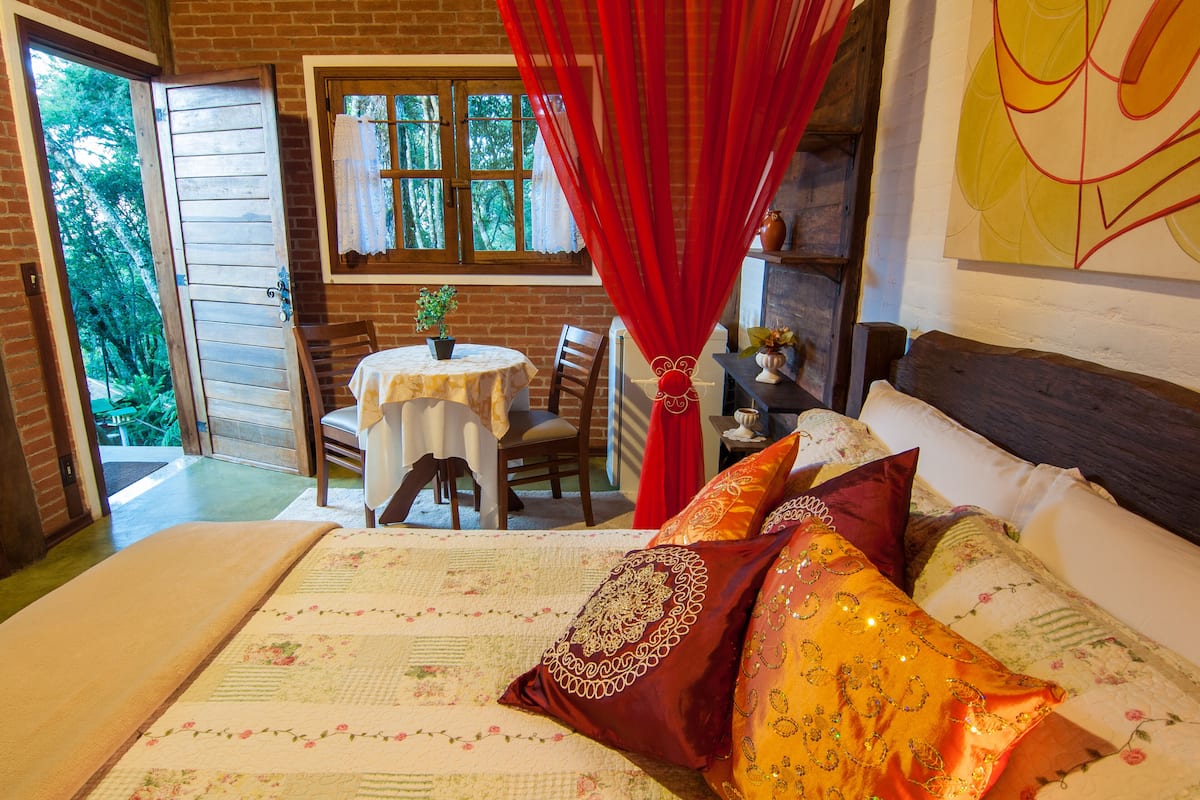 A bedroom with a bed, a window with curtains, a table with chairs, and a view of greenery outside.