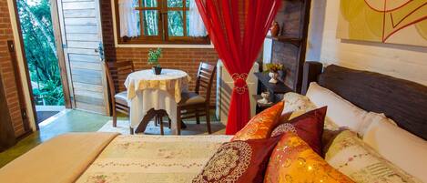 A bedroom with a bed, a window with curtains, a table with chairs, and a view of greenery outside.