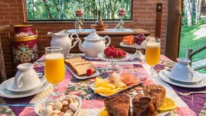 A breakfast table set with various foods and drinks, including bread, fruit, and a jug of orange juice, in a room with a brick wall and a window overlooking greenery. - Pousada Alto da Neblina (Camanducaia)