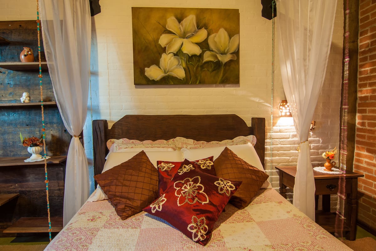 A bedroom with a bed, pillows, a nightstand, a vase with flowers, and a painting on the wall.