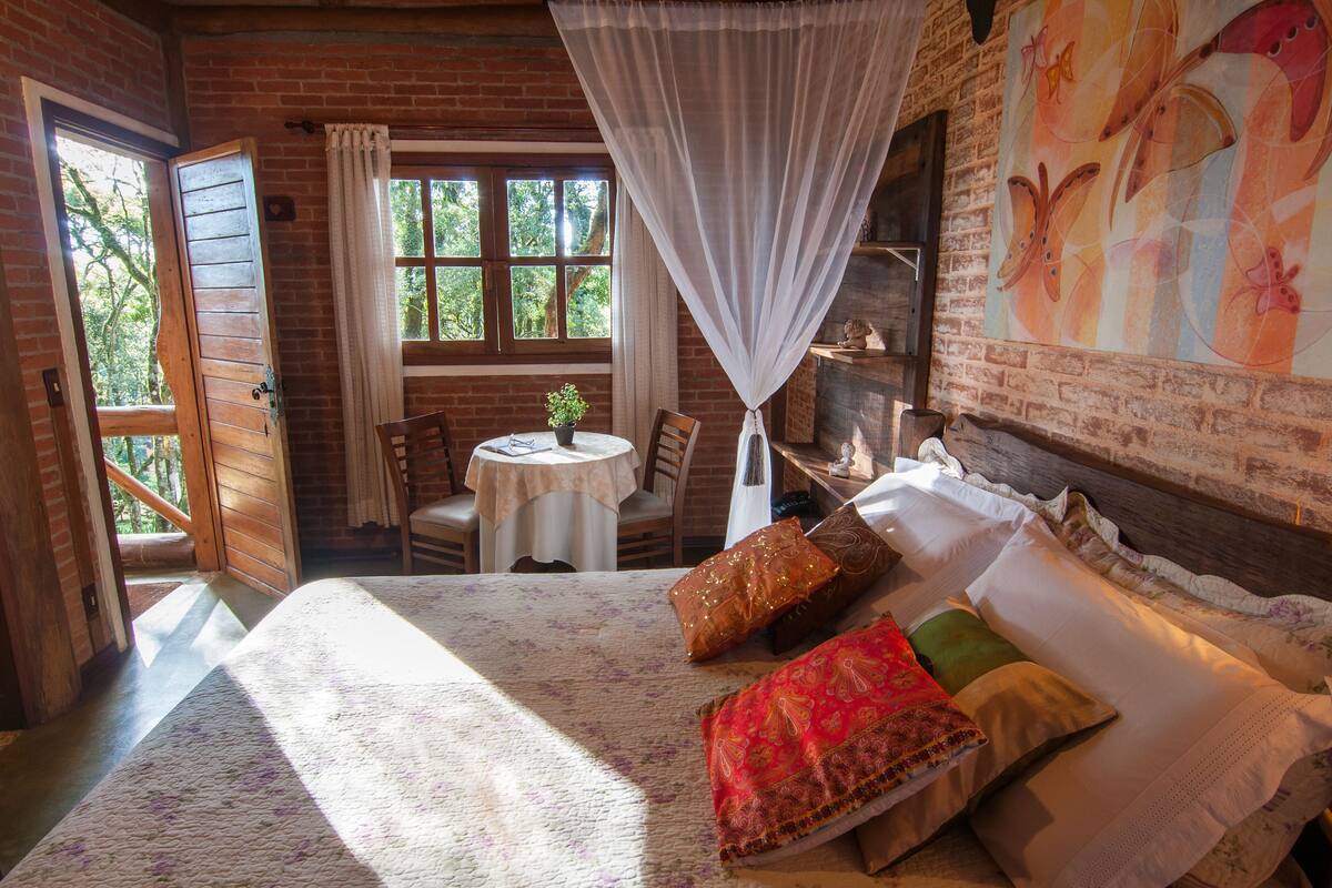 A room with a bed, a table, and a chair. There is a window with curtains and a view of trees.