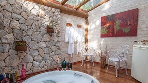 A spa room with a stone wall, a hot tub, white chairs, and a painting. - Pousada Alto da Neblina (Camanducaia)