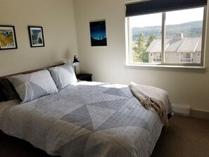 Un dormitorio con una cama grande, una mesita de noche, una lámpara y una ventana con vistas a casas y árboles.